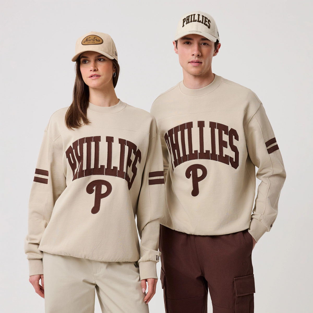 New Era Philadelphia Phillies MLB Arch Graphic Light Beige Crew Neck Sweater, front view on two models with matching light beige caps, showcasing large brown Phillies lettering and a P logo on the chest, complemented by double stripes on the sleeves.