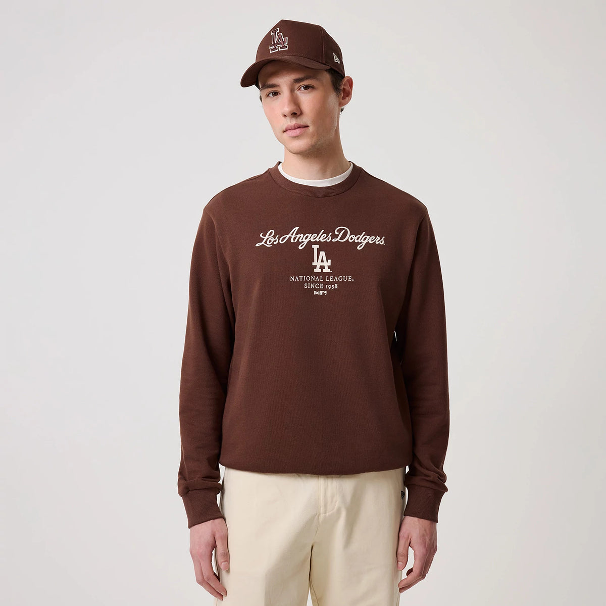 New Era LA Dodgers MLB Linear Brown Crew Neck Sweater, front view on a model wearing a matching brown cap, featuring white Los Angeles Dodgers script across the chest, the LA logo beneath, and ribbed cuffs and hem for a classic fit.