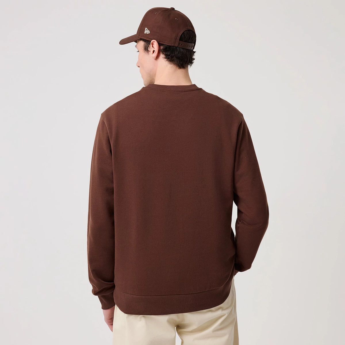 New Era LA Dodgers MLB Linear Brown Crew Neck Sweater, back view on a model wearing a matching brown cap, highlighting the solid brown fabric with ribbed cuffs and hem, creating a casual yet sporty look complemented by beige trousers.