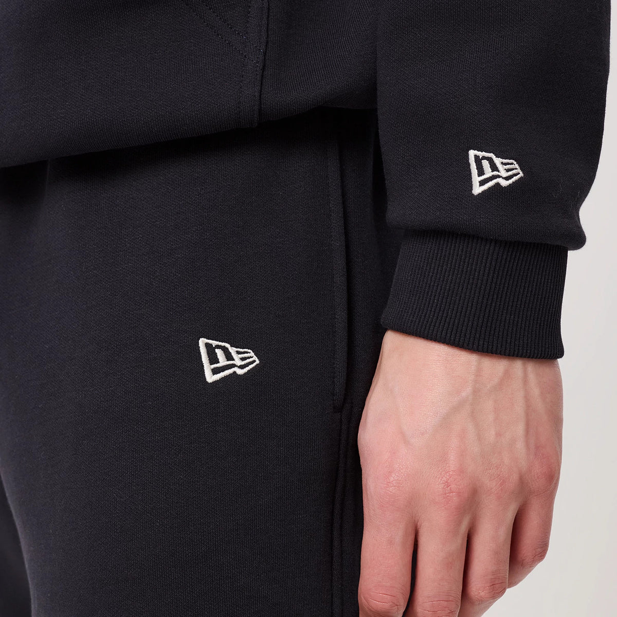 New Era Navy Straight Leg Joggers, close-up view showing the embroidered New Era flag logo in white on the pocket area and lower leg, with ribbed cuffs and an adjacent hand revealing the soft fabric texture.
