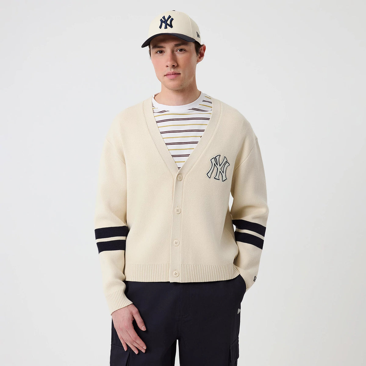 New Era New York Yankees MLB Knit Light Beige Cardigan, front view on a model styled with a matching cap, featuring the navy embroidered NY logo on the left chest, ribbed V-neckline, and contrasting black stripes on the sleeves.