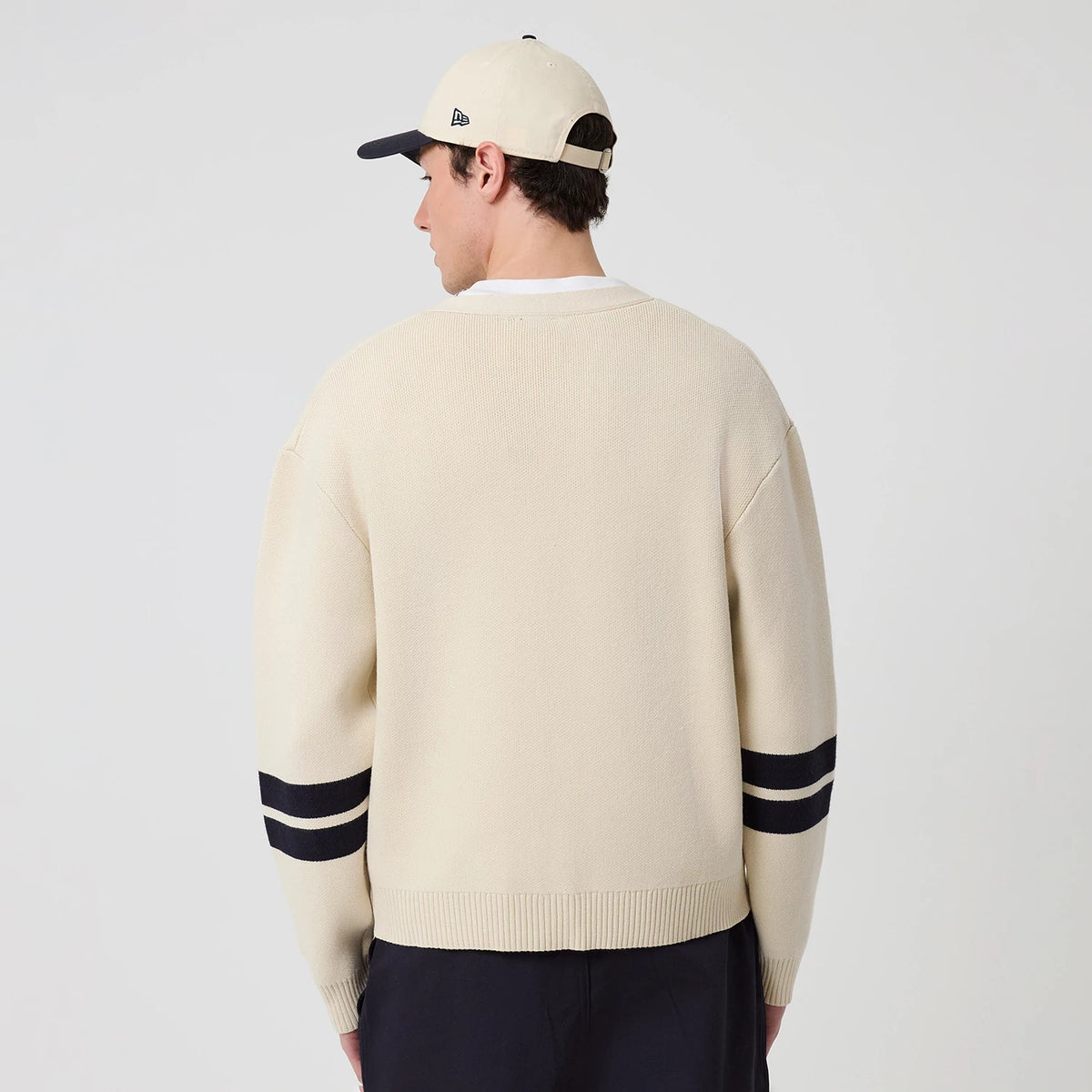 New Era New York Yankees MLB Knit Light Beige Cardigan, back view on a model showing the light beige knit fabric with black double stripes on the sleeves and a relaxed fit, complemented by a cream cap with the New Era flag logo on the side.