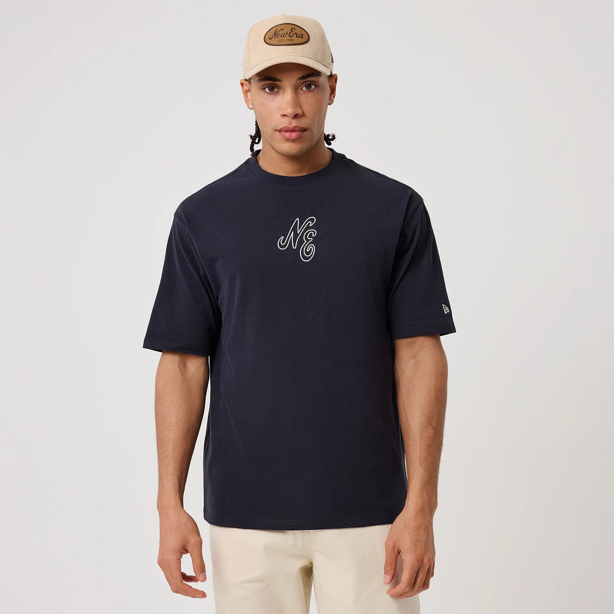 New Era Crest Graphic Navy Oversized T-Shirt, front view on a model, featuring a large embroidered NE logo centred on the chest, a small embroidered New Era flag logo on the left sleeve, and styled with a beige New Era cap.