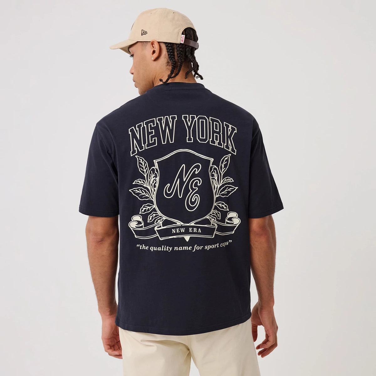 New Era Crest Graphic Navy Oversized T-Shirt, back view on a model with a beige cap, showing a large crest design with New York lettering at the top, the New Era shield emblem in the centre, and decorative leaves branching out.