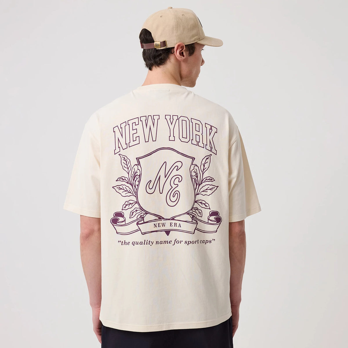 New Era Crest Graphic Open White Oversized T-Shirt, back view on a model styled with a beige cap, showing a New York graphic in purple, an ornate crest with NE initials, and the phrase the quality name for sport caps underneath.