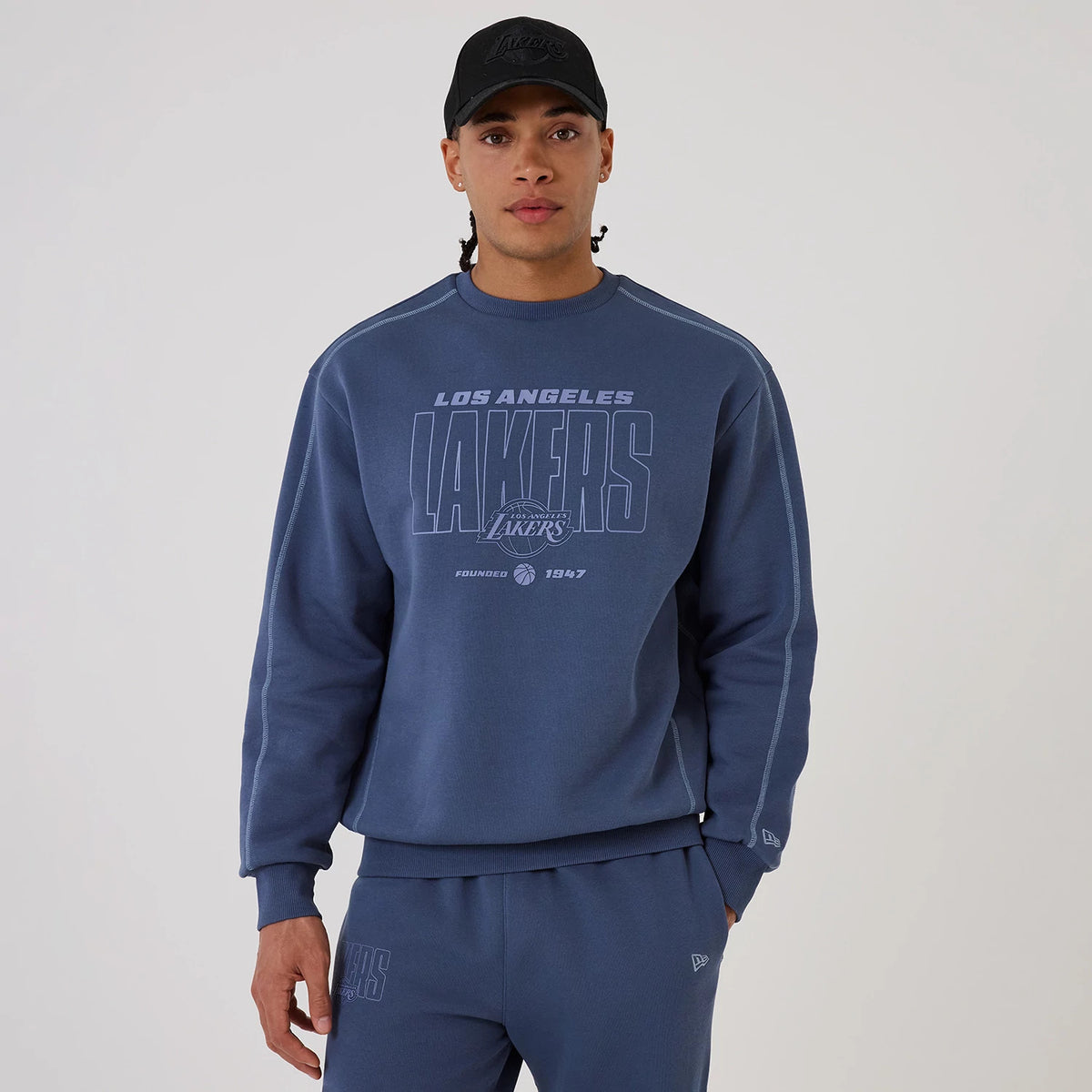 New Era LA Lakers NBA Graphic Dark Blue Crew Neck Sweater, front view on a model wearing matching dark blue track pants, showcasing a large LAKERS graphic with team logo across the chest, complemented by ribbed cuffs and waistband.