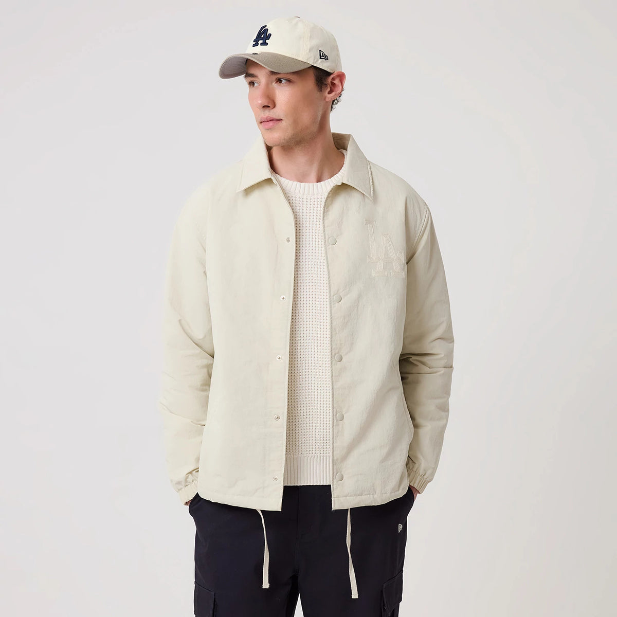 New Era LA Dodgers MLB Light Beige Coach Jacket, front view on a model styled with a light beige cap, showcasing an embroidered LA Dodgers logo on the left chest, button fastening down the centre, and drawstrings hanging from the hemline.
