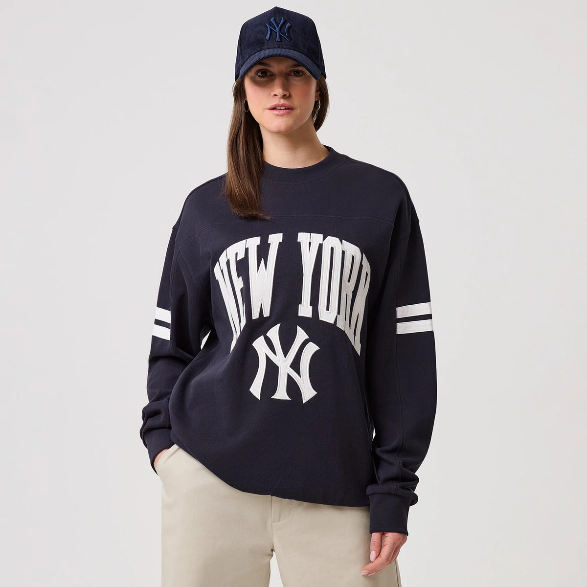 New Era New York Yankees MLB Arch Graphic Navy Crew Neck Sweater, front view on a model with a matching navy cap, featuring white New York lettering across the chest, the NY logo beneath, and double white stripes on each sleeve.