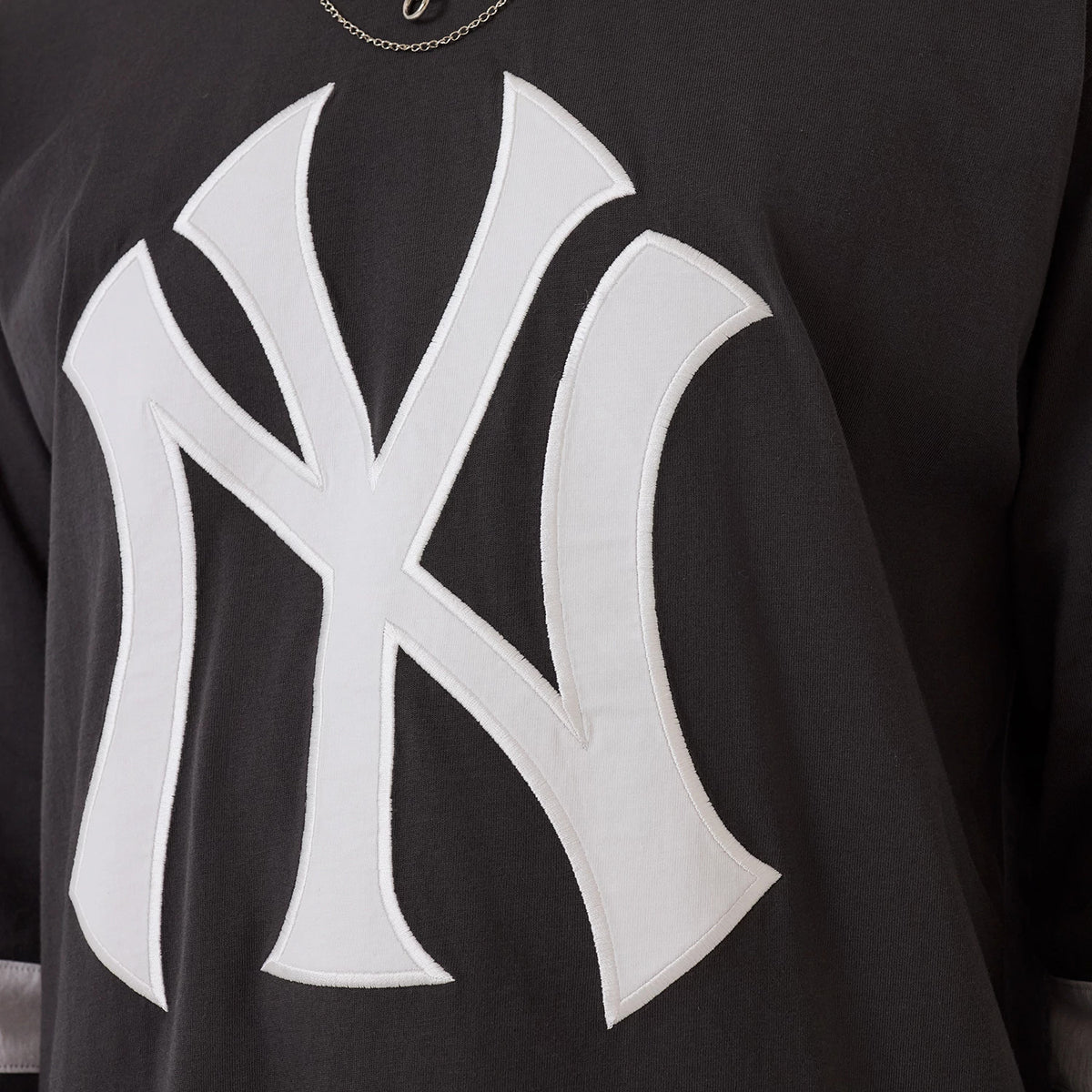 New Era New York Yankees MLB League Essential Charcoal Oversized Jersey, front view showing a large white embroidered NY logo on the charcoal fabric, with stitching around the edges and a glimpse of a silver chain necklace above.