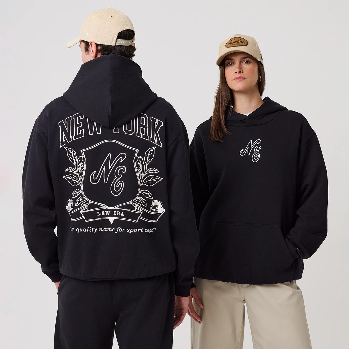 New Era Crest Graphic Navy Pullover Hoodie, front and back view on models showing a large white printed crest and New York lettering on the back, with a smaller NE logo on the chest, both models wearing beige caps for a casual style.