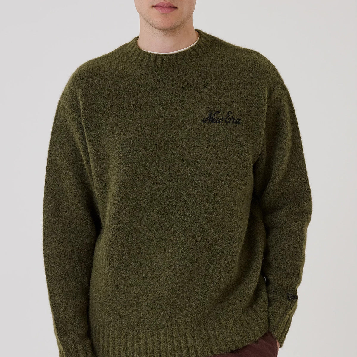New Era Simple Knit Green Knitted Sweater, front view on a model wearing brown trousers, featuring black embroidered New Era branding on the left chest, ribbed crew neckline, and textured knitted fabric with a warm, casual appearance.