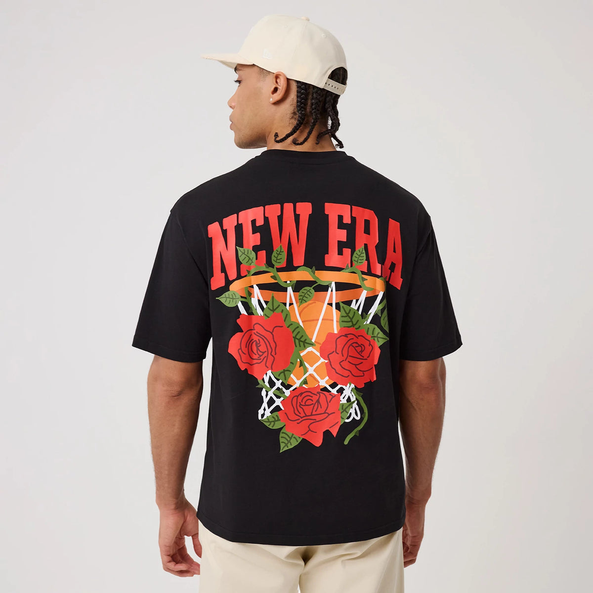 New Era Floral Graphic Black T-Shirt, back view on-model displaying red New Era lettering at the top, a large graphic of roses and a basketball hoop below, paired with a neutral cap and light trousers for a casual look.