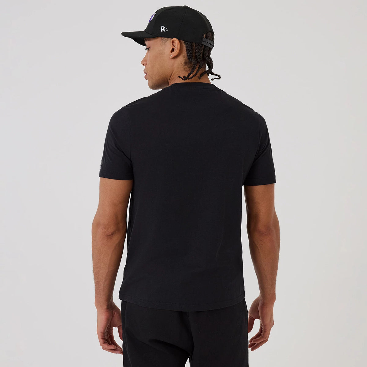 New Era LA Lakers NBA Graphic Black T-Shirt, back view on-model showing plain black fabric with a small, embroidered New Era logo on the left sleeve, styled with black trousers and a black cap featuring an embroidered New Era logo on the side.