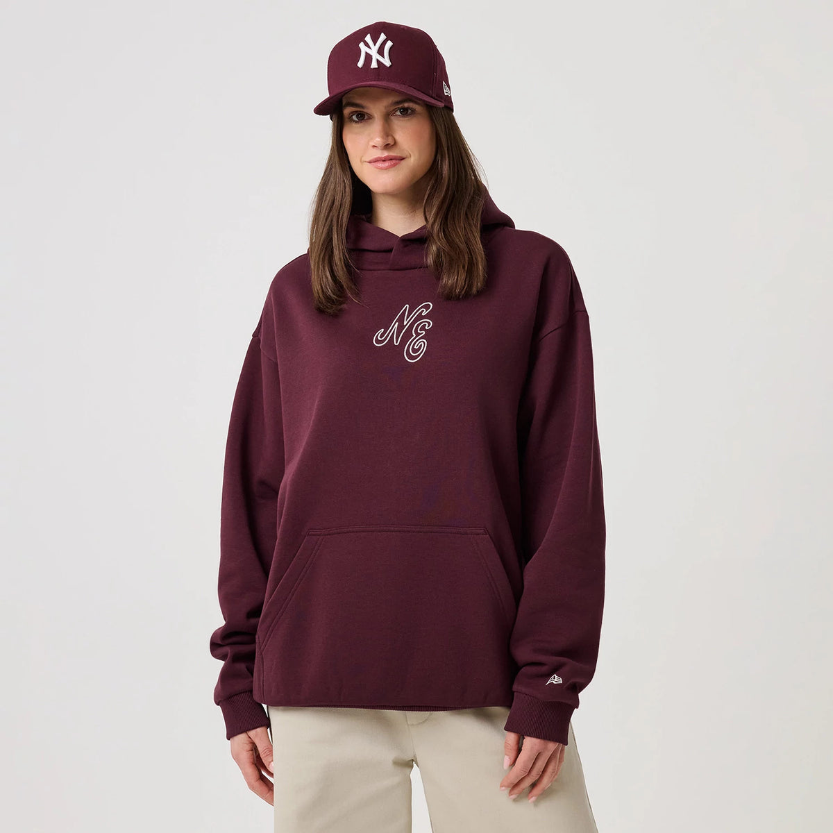 New Era Crest Graphic Dark Purple Pullover Hoodie, front view on a model styled with a matching dark purple cap, showing an embroidered NE logo on the centre chest, ribbed cuffs, and a small embroidered New Era logo on the sleeve.
