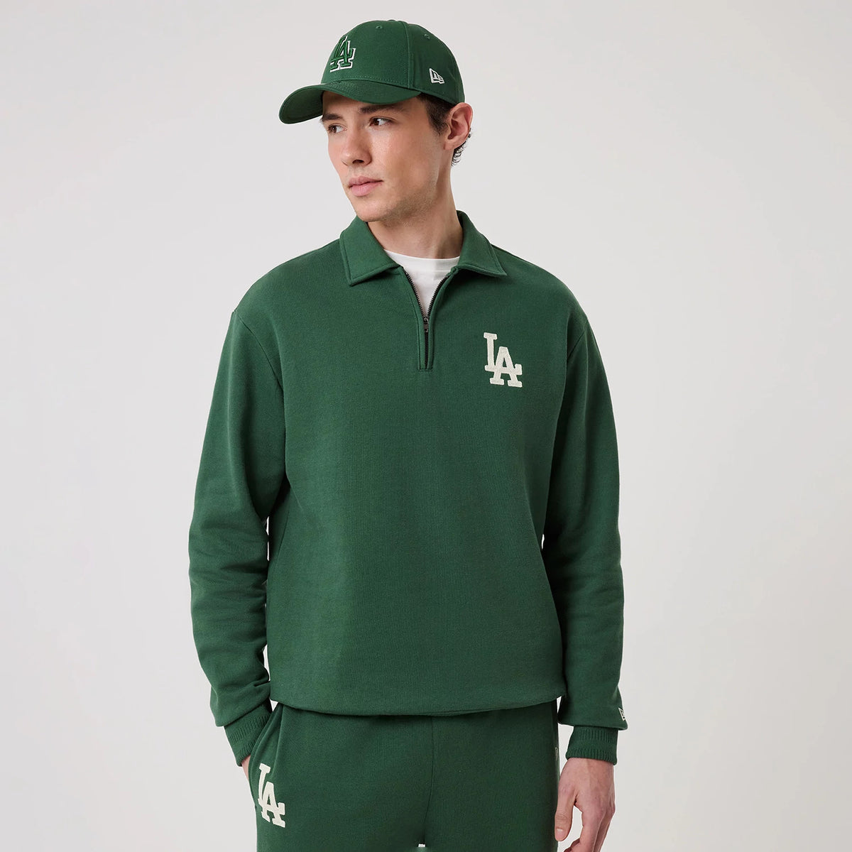 New Era LA Dodgers MLB Dark Green 1/4 Zip Sweater, front view on a model wearing a matching cap and joggers, showing a white LA logo on the left chest, ribbed cuffs, and a quarter-zip design with a stand-up collar.