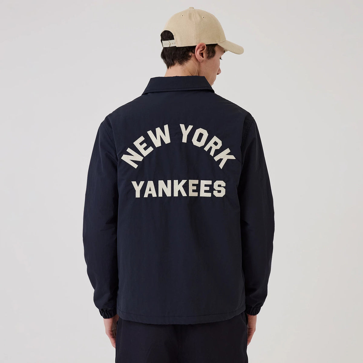New Era New York Yankees MLB Navy Coach Jacket, back view on-model showing large white embroidered New York Yankees lettering across the upper back, a classic collared design, and elasticated cuffs, paired with a beige cap for a casual look.