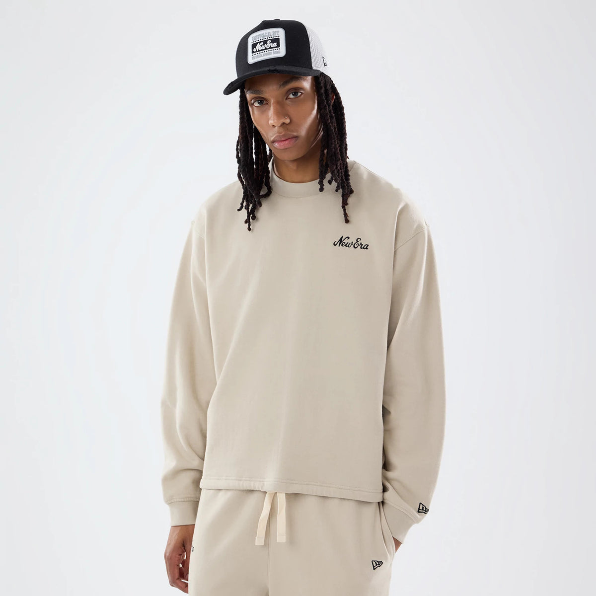 New Era Open Hem Light Beige Oversized Crew Neck Sweater, front view on a model wearing a black cap, featuring an embroidered black New Era script logo on the left chest and the New Era flag logo on the left cuff.
