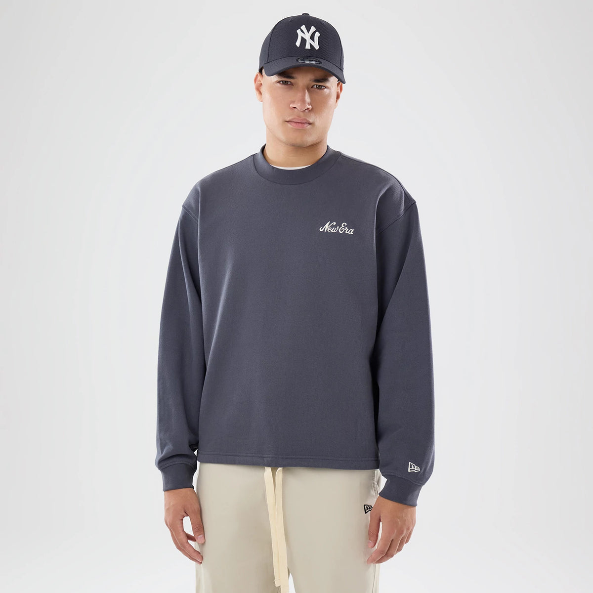 New Era Open Hem Dark Grey Oversized Crew Neck Sweater, front view on a model wearing a dark cap with a white logo, showing the white embroidered New Era script on the chest and the New Era flag logo on the left sleeve.