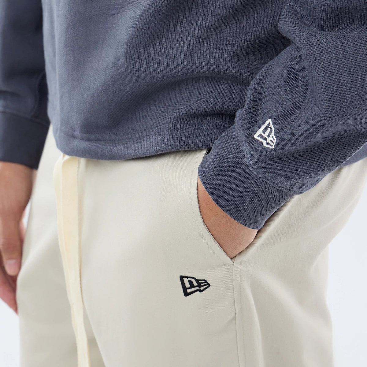 New Era Open Hem Dark Grey Oversized Crew Neck Sweater, on-model view showing the left cuff with an embroidered white New Era flag logo, relaxed fit drape over off-white trousers, and a straight hemline just above the waistband.