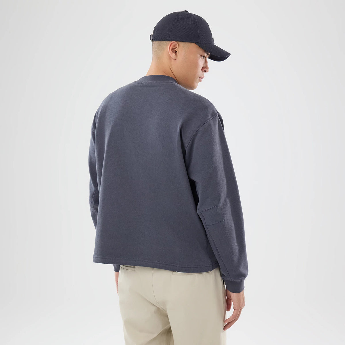 New Era Open Hem Dark Grey Oversized Crew Neck Sweater, back view on a model paired with a dark cap, showing the relaxed fit and drop shoulder design of the dark grey sweater, complemented by light-coloured trousers.
