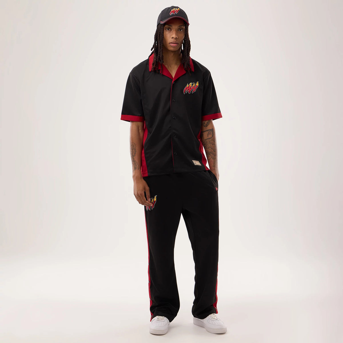 New Era Miami Heat NBA Hardwood Classic Straight Black Straight Leg Joggers, front view on a model with black joggers featuring an embroidered Miami Heat flame logo on the left thigh, red side panels, and a relaxed fit down to white trainers.