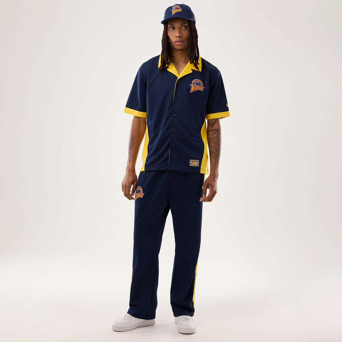 New Era Warriors NBA Hardwood Classic Revere Dark Blue Short Sleeve Shirt, front view on a model with a matching cap, showcasing a team logo on the left chest, contrasting yellow collar and trim, and button-front style.
