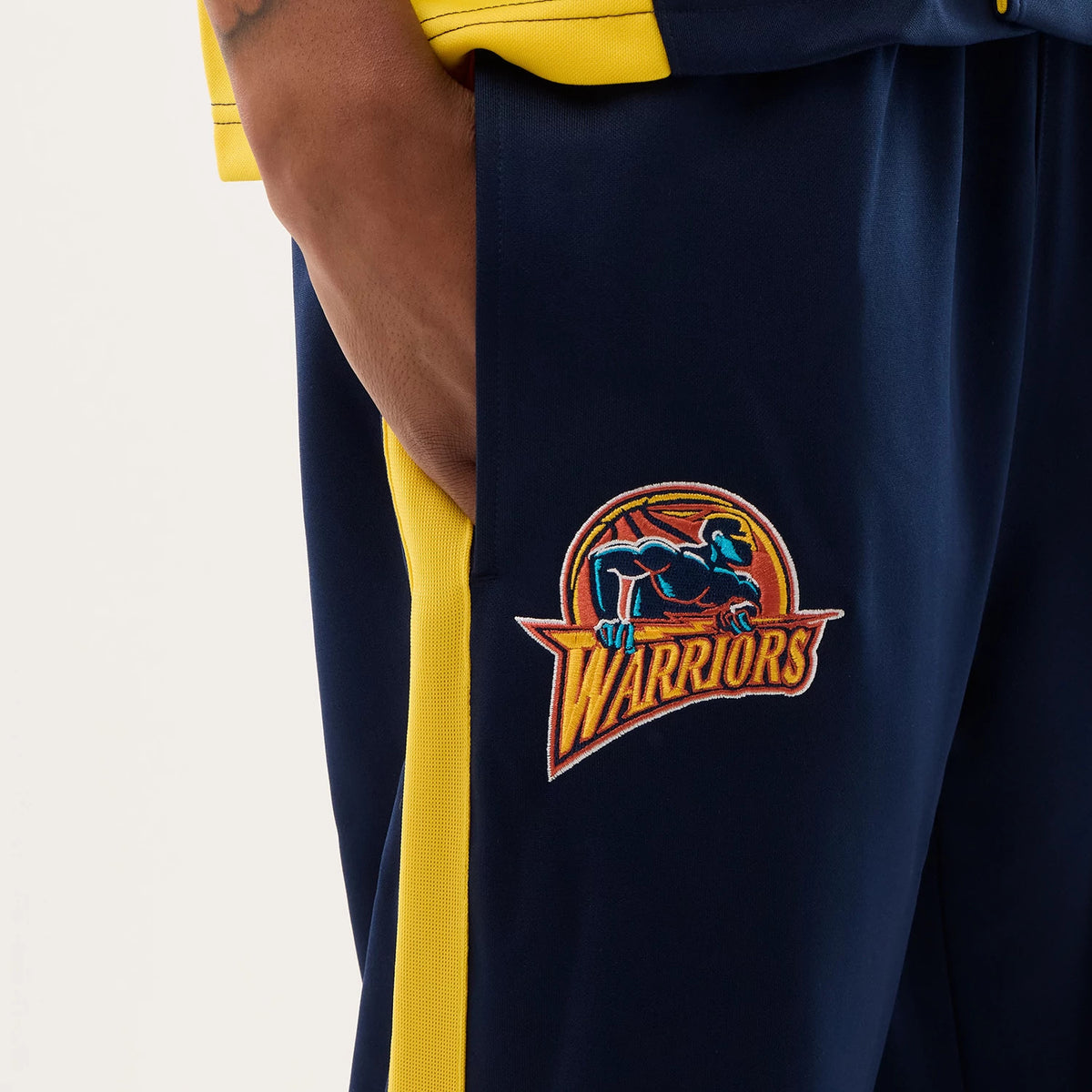 New Era Warriors NBA Hardwood Classic Straight Dark Blue Straight Leg Joggers, side view showing the embroidered Warriors logo on the right thigh with a detailed blue figure and red basketball design, contrasting with yellow side stripes.