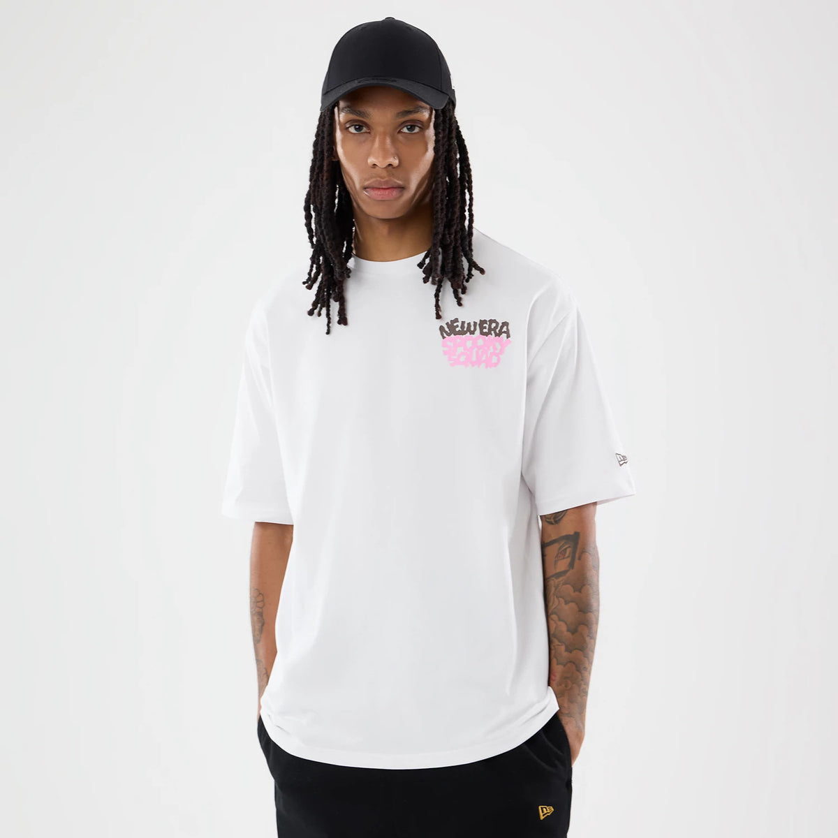 New Era Spooky Graphic Oversized White T-Shirt, front view on-model showing a pink graffiti-style graphic and brown New Era lettering on the chest, a embroidered New Era logo on the left sleeve, and styled with a black cap.