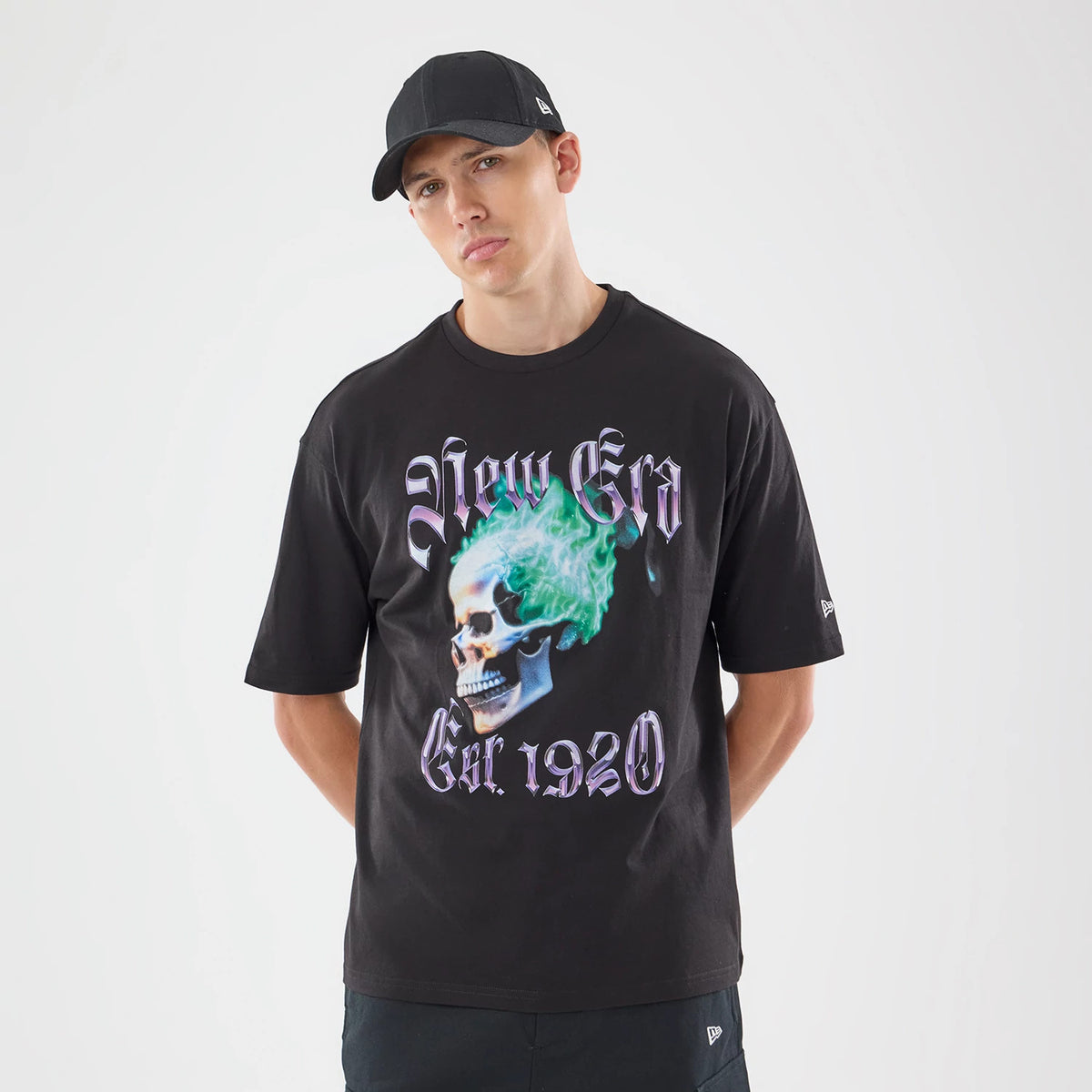 New Era Skull Graphic Oversized Black T-Shirt, front view on a model with a black cap, featuring a skull graphic with green flames centred on the chest, purple New Era Est. 1920 lettering, and an embroidered New Era logo on the sleeve.
