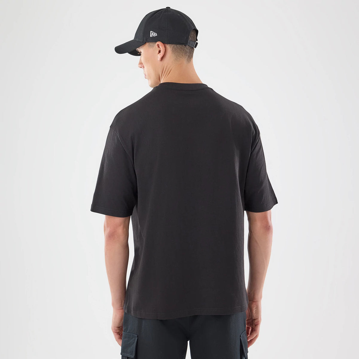 New Era Skull Graphic Oversized Black T-Shirt, back view on a model wearing a black cap, showing the plain black fabric of the oversized tee without graphics, loose short sleeves and a relaxed fit over dark trousers.