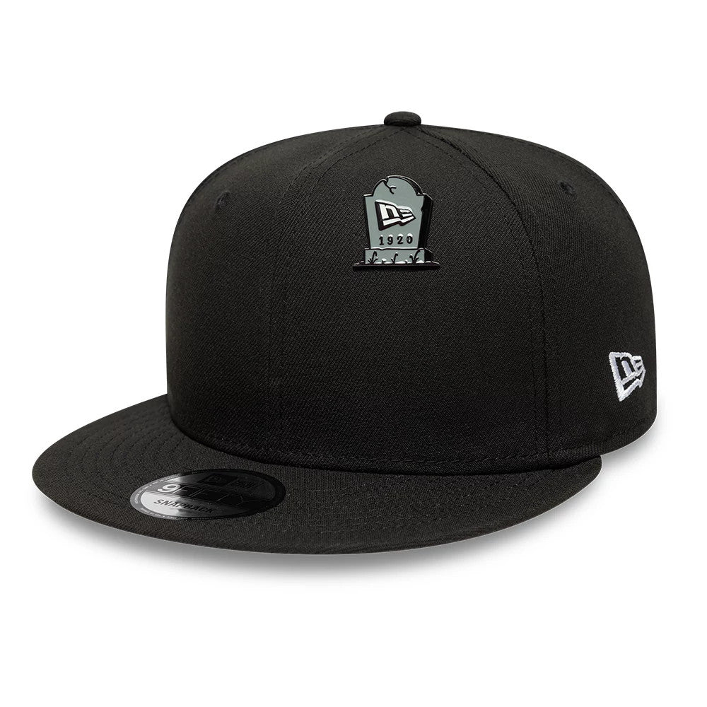 New Era Halloween Grave Grey Pin Badge, three-quarter view showing a black cap with a grey tombstone-shaped pin badge featuring the New Era logo and 1920 on the front, an embroidered New Era flag logo on the side, and a flat black visor.