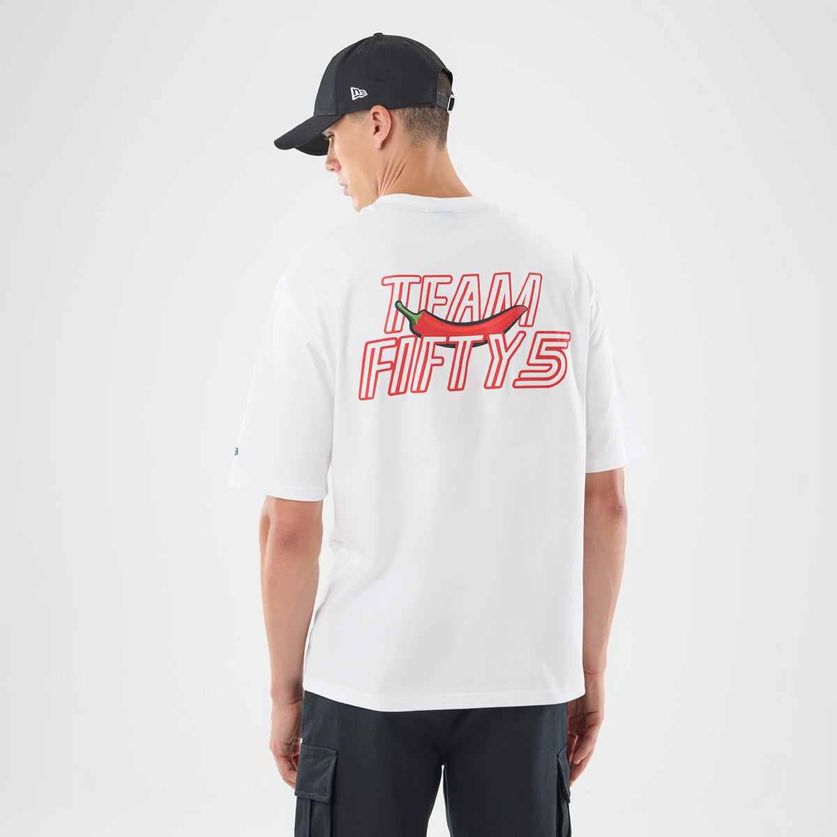 New Era Carlos Sainz Team Fifty5 Mexico White Oversized T-Shirt, back view on a model showing red and white Team Fifty5 text across the shoulders with a red chilli pepper graphic beneath, styled with loose black cargo trousers and a black cap.