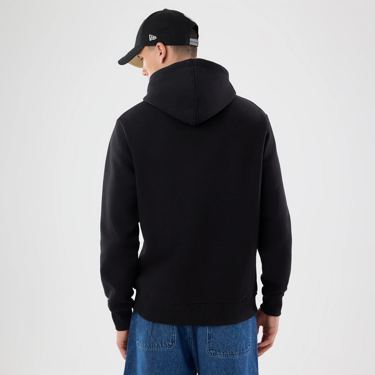 New Era New Orleans Saints NFL Patch Black Pullover Hoodie, back view on a model wearing relaxed jeans and a matching black cap, showing a plain black design with the hood up and no graphics or logos on the back.