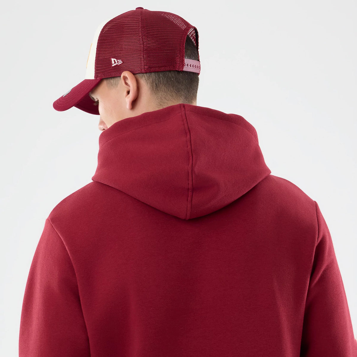 New Era Washington Commanders NFL Patch Dark Red Pullover Hoodie, back view on a model wearing a matching dark red mesh cap, showcasing the large hood and seamless tailored design of the dark red fabric extending over the shoulders and upper back.