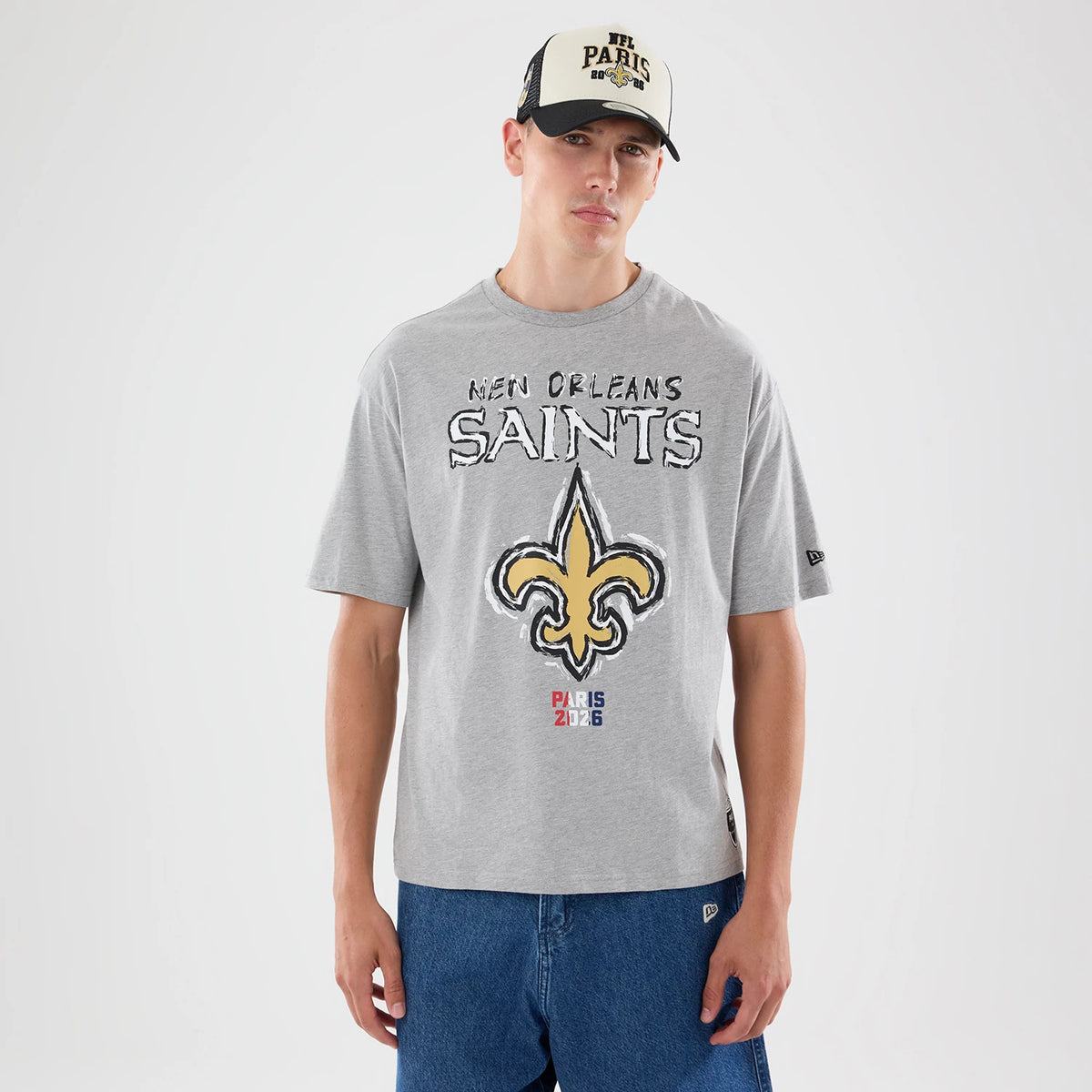 New Era New Orleans Saints NFL Graphic Slogan Grey Oversized T-Shirt, front view on a model styled with a black and white cap, showcasing the large central fleur-de-lis graphic beneath New Orleans Saints lettering and Paris 2023 text at the bottom.