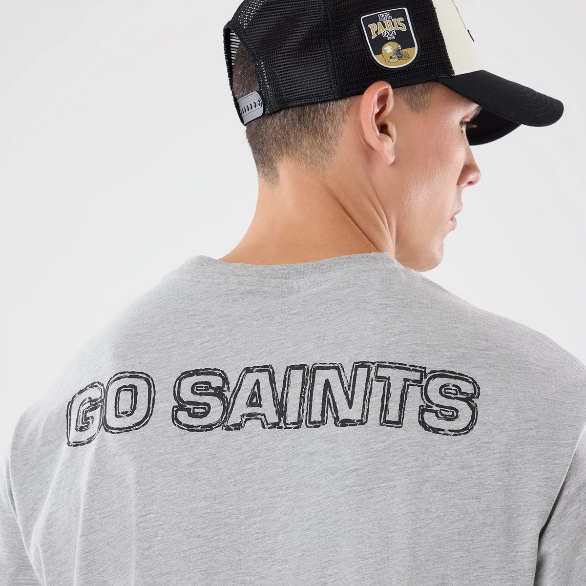 New Era New Orleans Saints NFL Graphic Slogan Grey Oversized T-Shirt, back view on-model showing black GO SAINTS lettering across the upper back, styled with a mesh trucker cap featuring a raised New Orleans Saints patch on the side.