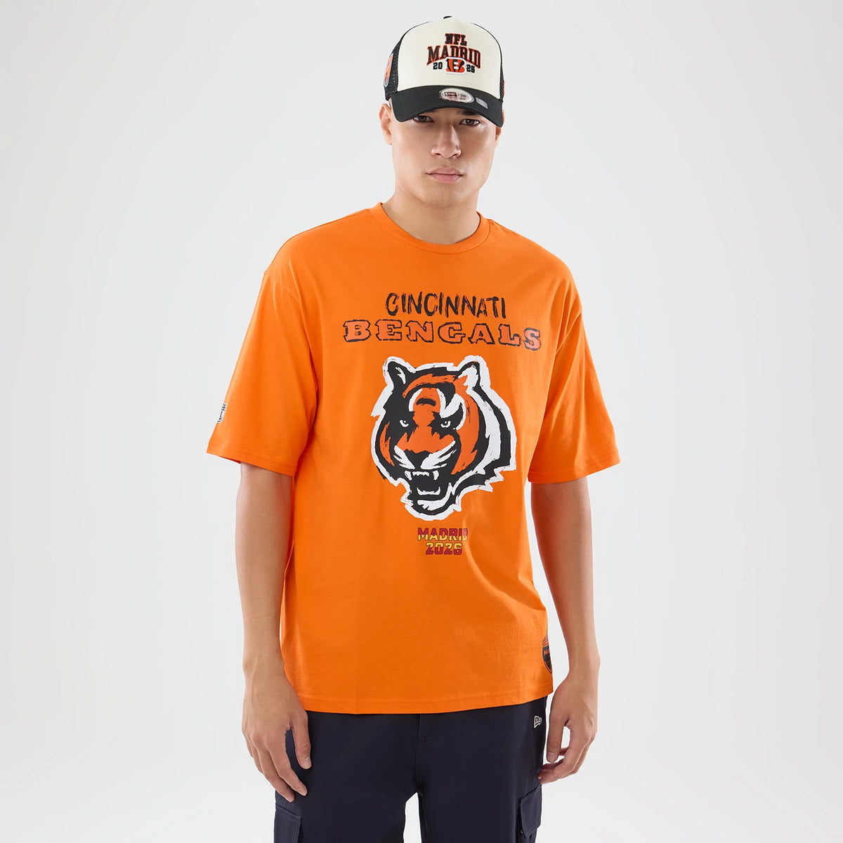 New Era Cincinnati Bengals NFL International Games 2026 Orange Oversized T-Shirt, front view on a model wearing a matching cap, with the Cincinnati Bengals text above a fierce tiger face graphic and Madrid 2026 details prominently below the logo.