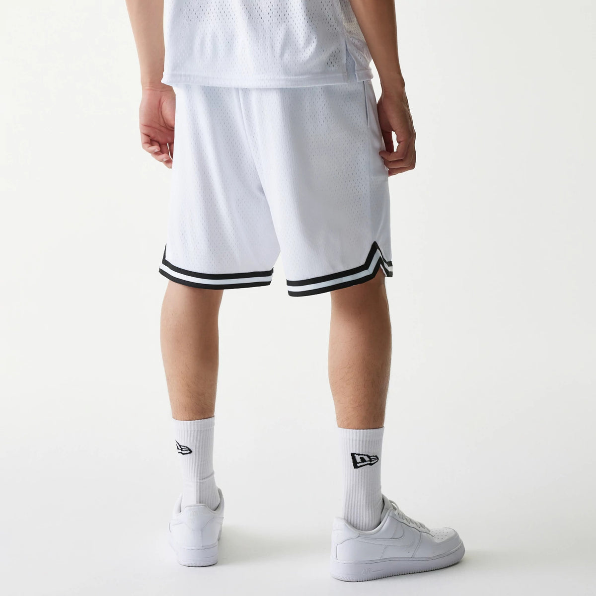 New Era Japan Mesh White Shorts, rear view showing the loose fit mesh fabric design, black and white striped trim at the hem, and the shorts falling just above the knees, styled with matching white socks and trainers.