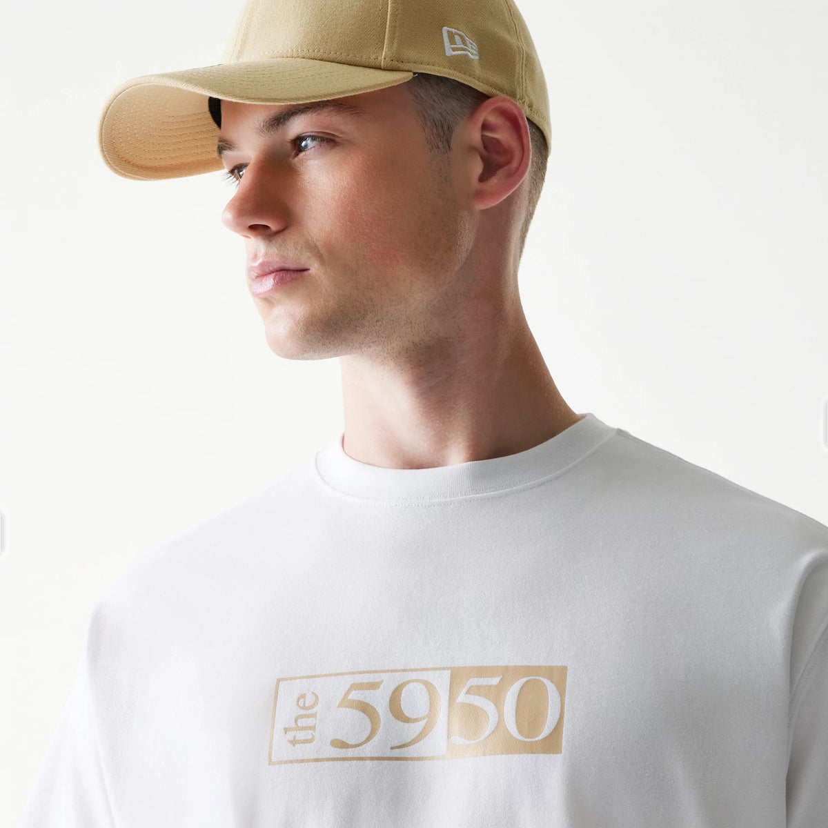 59FIFTY Fitted Cap Description Graphic White T-Shirt, on-model view showing the front graphic in beige letters across the chest, a person in a beige cap with a debossed New Era logo on the side wearing the shirt.