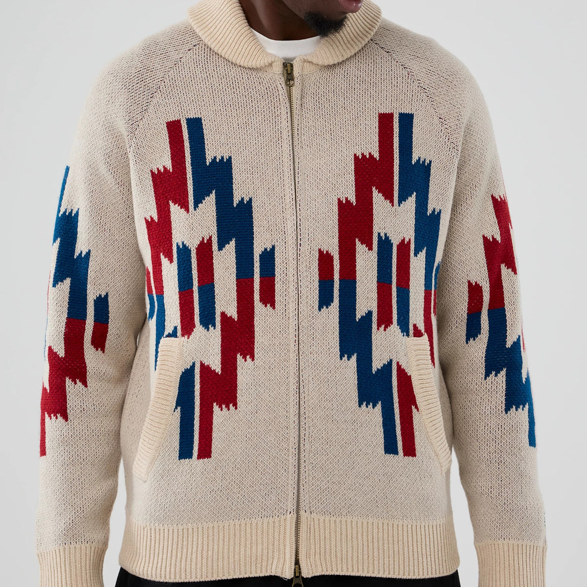 New Era Organic Cowichan Natural Cardigan, front view on a model showing a zip closure and intricate red and blue geometric patterns across the chest and sleeves, with a ribbed collar and hem.