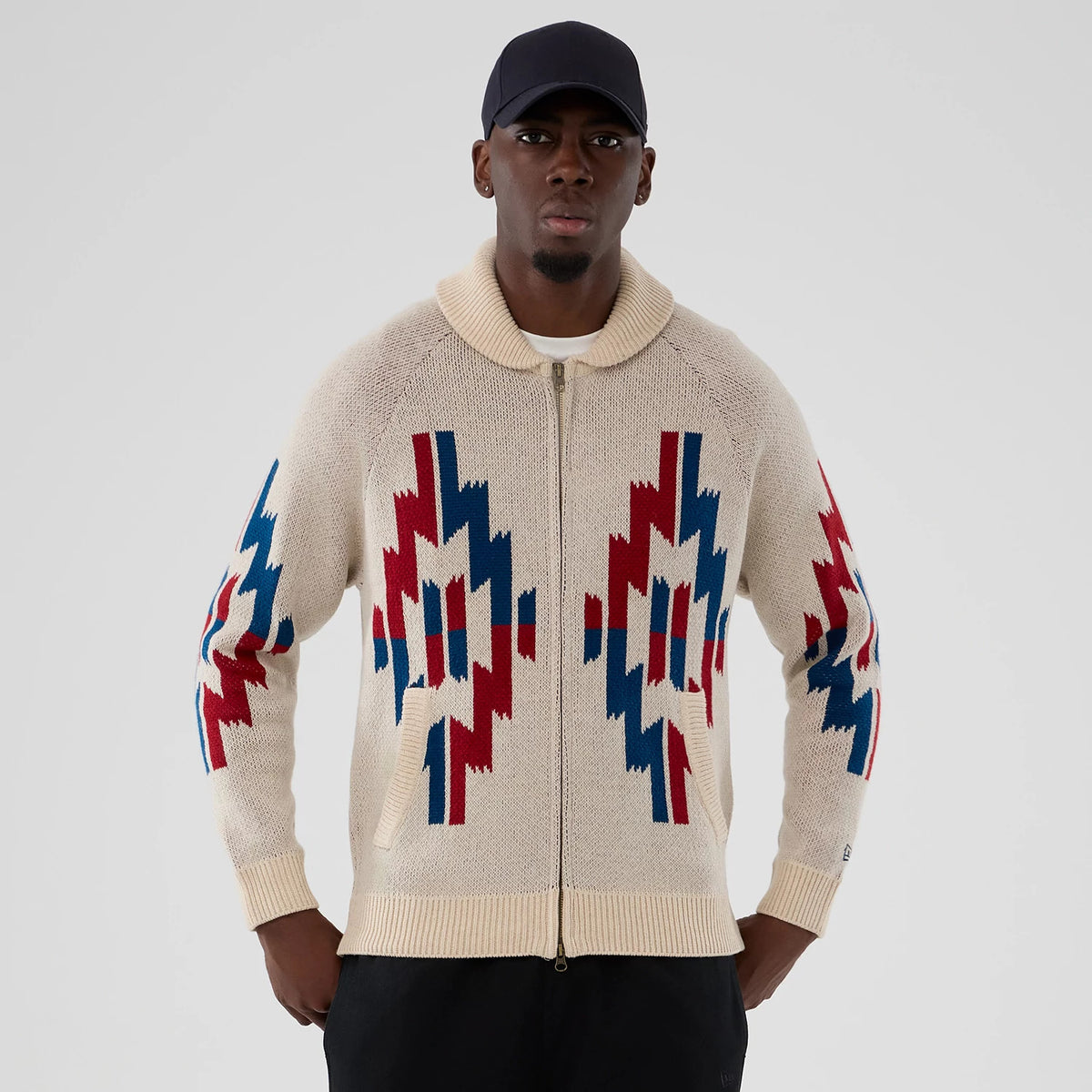 New Era Organic Cowichan Natural Cardigan, front view on a model wearing a dark cap, featuring a zip-up design with vibrant red and blue geometric patterns on the sleeves and front, and ribbed cuffs and hem.