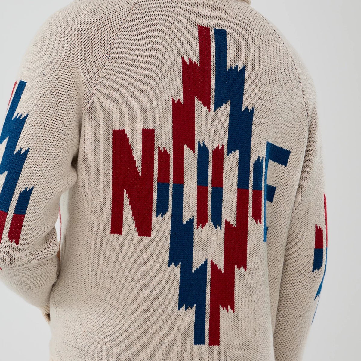 New Era Organic Cowichan Natural Cardigan, back view showing a large geometric pattern in red and blue across the back, with N logo elements on the sleeves, knitted in a natural beige fabric.
