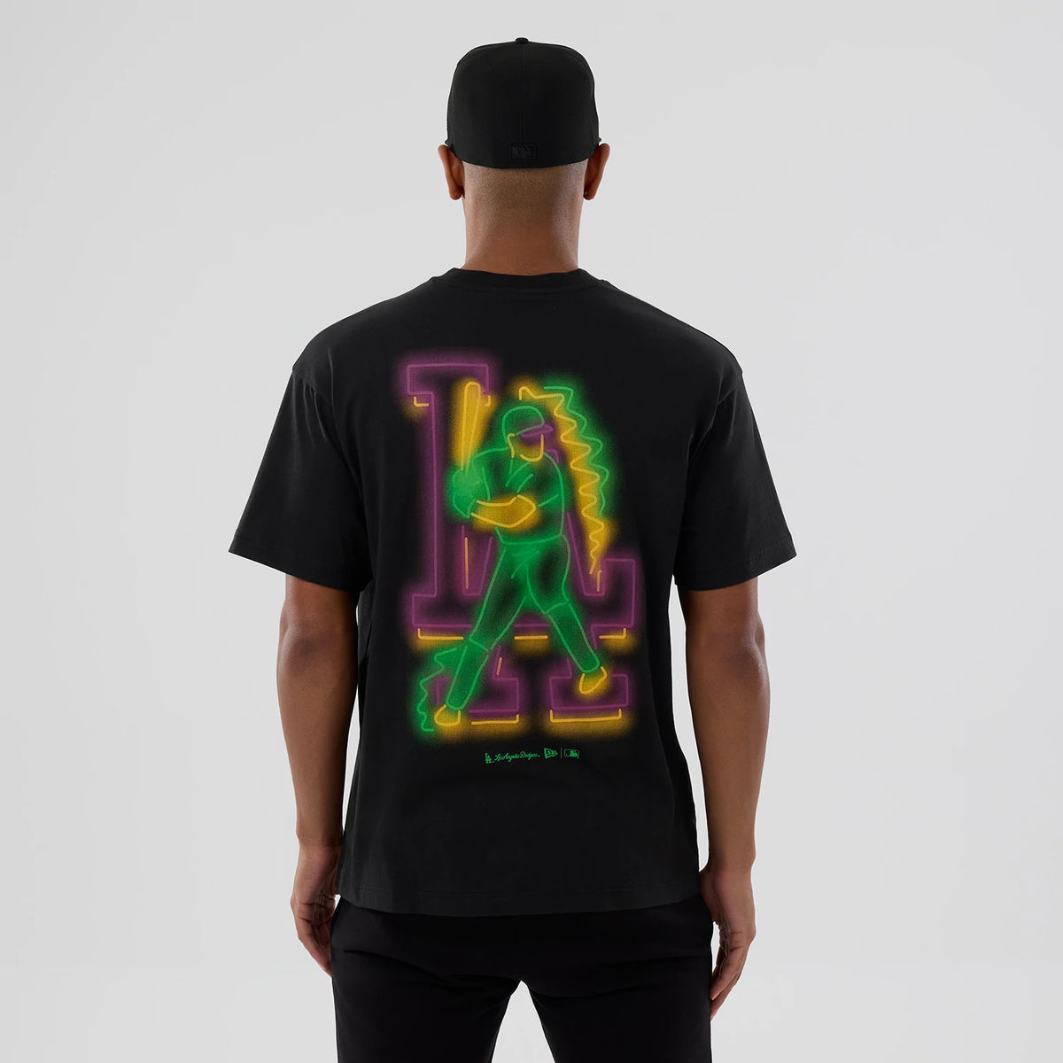 LA Dodgers MLB Neon Light Black T-Shirt, rear view showing a vibrant neon graphic of a baseball player in green and yellow over purple LA initials, worn by a person also wearing a black cap.