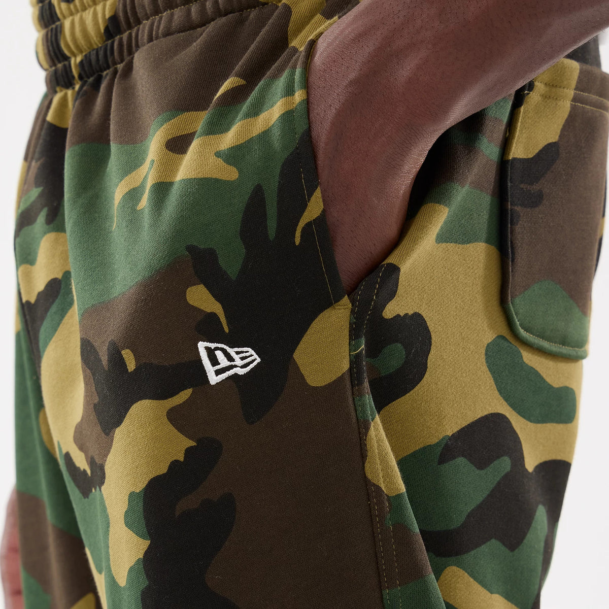 New Era Woodland Camo Joggers, close-up showing a side pocket and white New Era flag logo embroidered on camouflage fabric, with an elasticated waistband and a hand resting casually in the pocket.