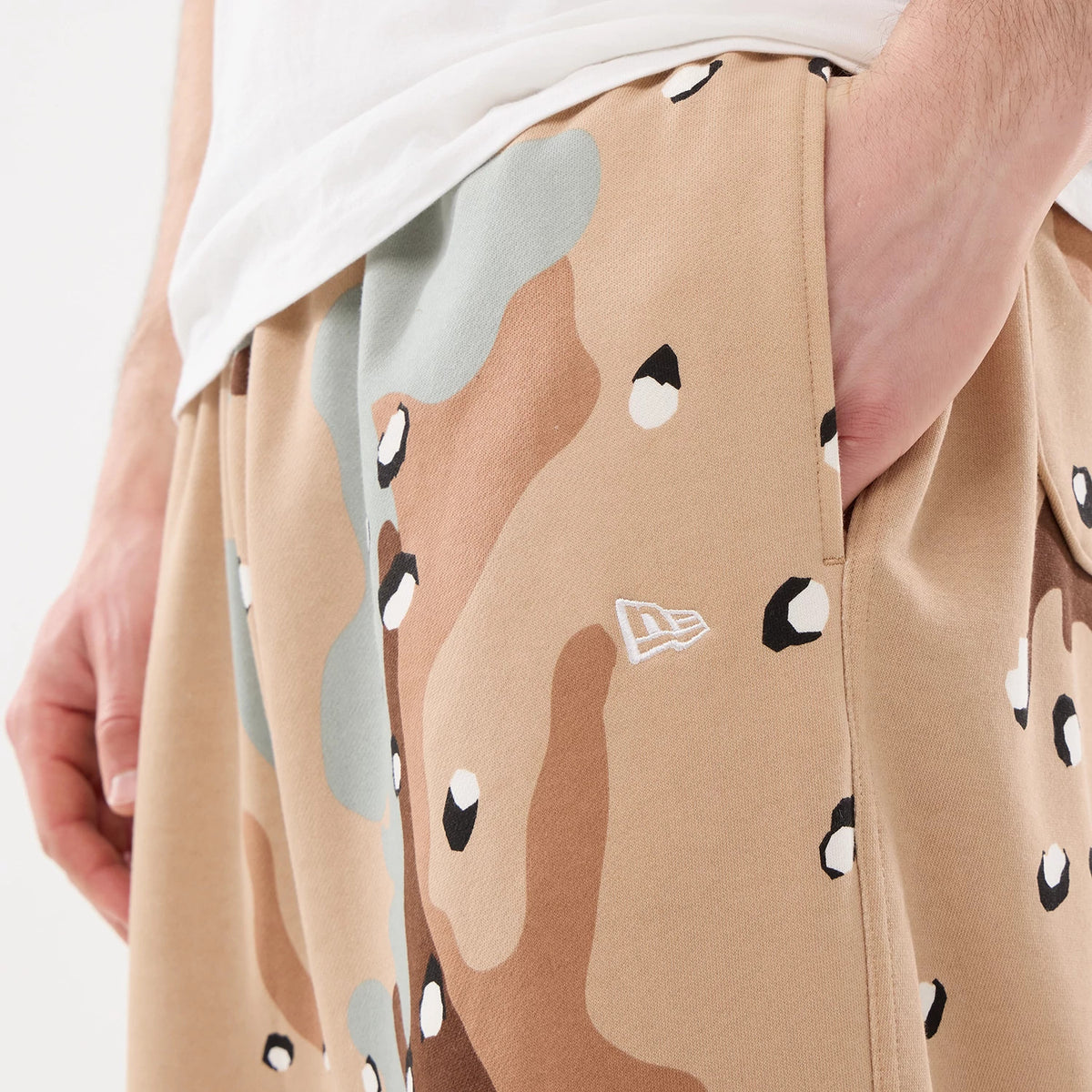 New Era Desert Camo Joggers, side view with a hand in the pocket, showing the distinctive desert camouflage pattern with contrasting shapes and the white embroidered New Era flag logo above the left pocket.