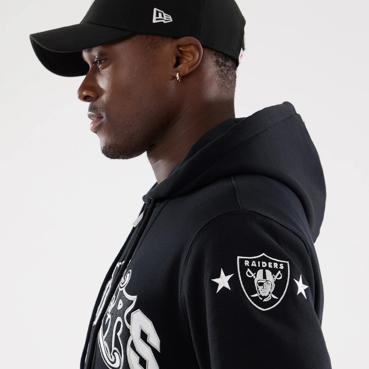 Las Vegas Raiders NFL Word Art Black 2 Way Full Zip Hoodie, side view showing a raised Raiders emblem on the sleeve and bold lettering across the chest, with a black New Era cap featuring a raised logo on the side.