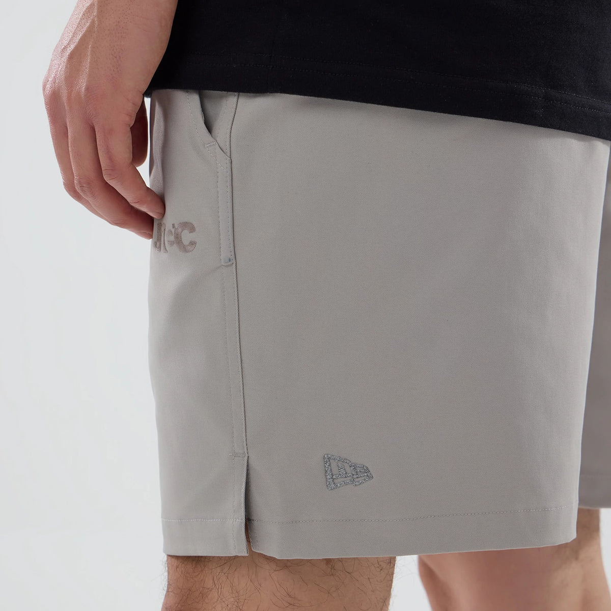 New Era NEC Logo Beige Oversized Shorts, side view showing the light beige fabric with a raised NEC logo near the pocket, a small raised New Era logo near the hem, and straight side seams.