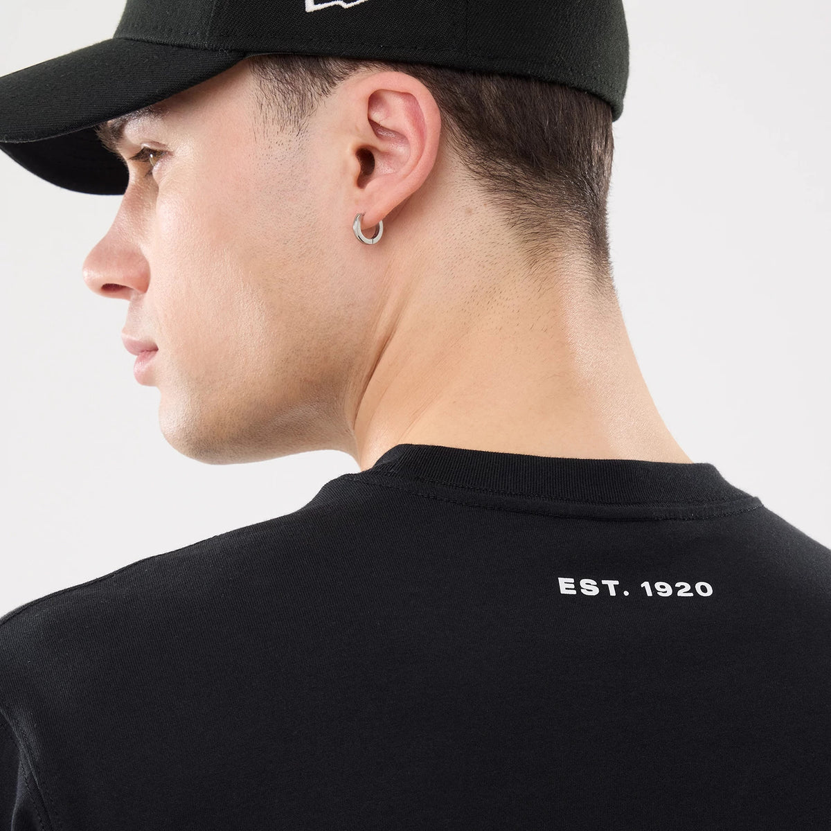 New Era Outdoor Mountain Black T-Shirt, on-model view showing the back of a black T-shirt with "EST. 1920" printed in white near the neckline, worn by a person in a black cap with a raised logo on the side.
