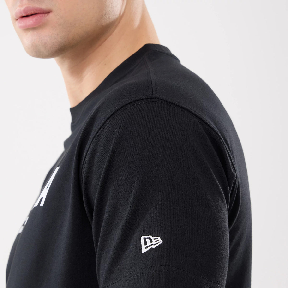 New Era Basic Black T-Shirt, side view showing the black sleeve with a raised New Era logo in white embroidery near the hem and a portion of the upper chest with a seamless shoulder seam.