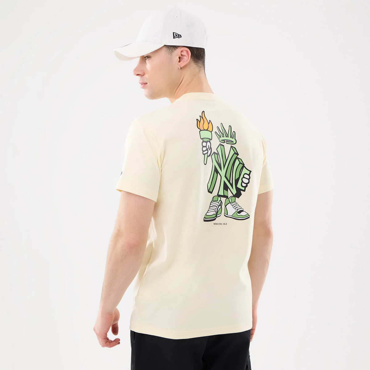 New York Yankees MLB Cartoon Cream T-Shirt, rear view showing a cartoon graphic of a Statue of Liberty character in Yankee-themed attire on the back, worn by a model in a white cap with a raised New Era logo on the side.